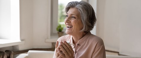 60 år gammel dame smiler i sofaen og holder rundt en tekopp. 